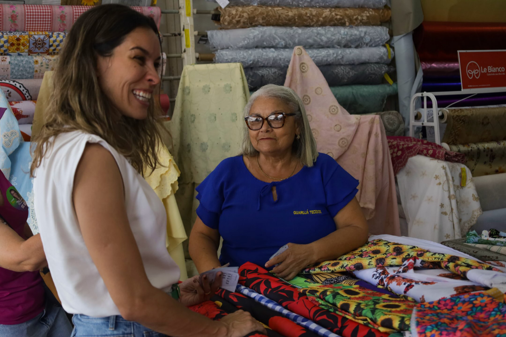 Mariana Dantas sorrindo ao lado de trabalhadora em banca de tecidos